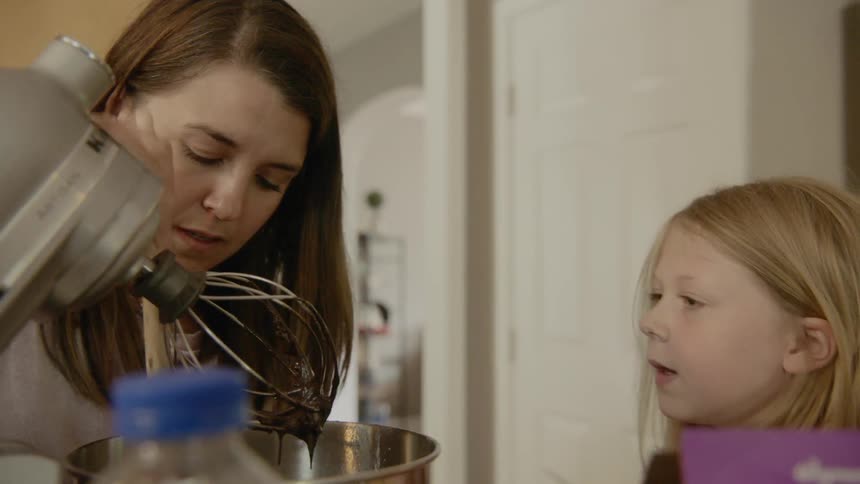 A young girl mixes frosting in a mixing bowl with her mother