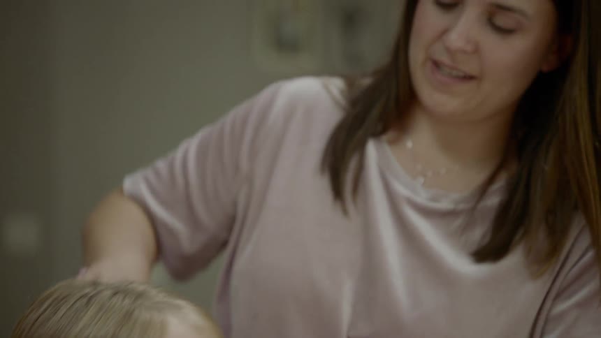 A young girl with straight blonde hair has it brushed by her mother as the two of them smile while the mother talks