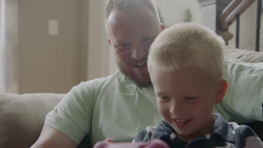 A man sits on the couch with a little boy in his lap playing on an electronic device