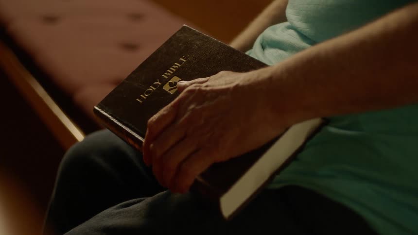 A person sits on a church pew while holding a bible