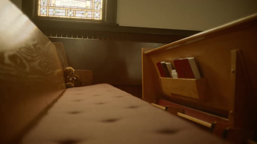A teddy bear sits at the end of an empty church pew under a stained glass window