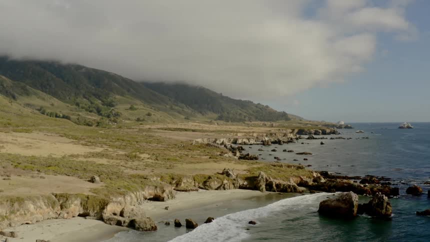 Waves come to a rocky shore with clouds hanging over the peaks and ridge of green hills