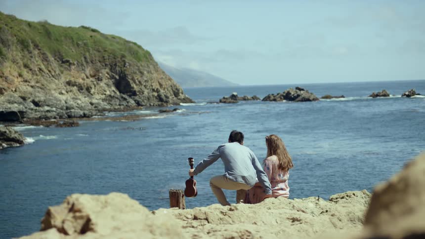 A man and woman sit by the water with a small guitar