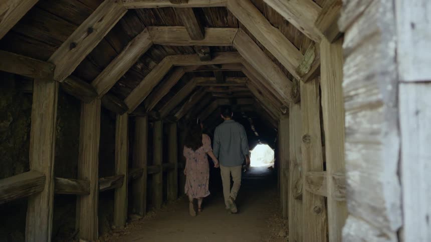 A couple walk hand in hand through a covered bridge while the man holds a ukulele