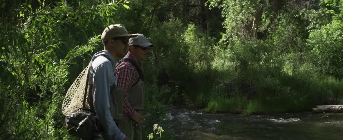 Two men with fishing gear standing next to a river surrounded by trees and brush on a chilly, sunny day
