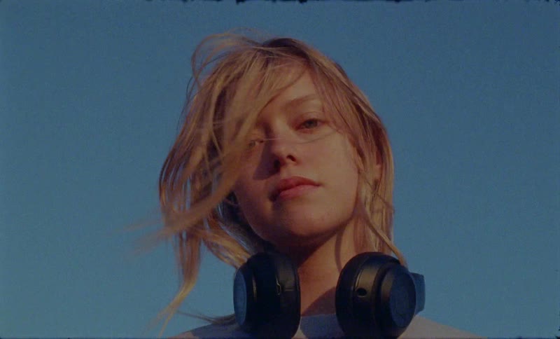 A young blonde female takes a pair of headphones off her ears and stands outside looking at the camera on a windy day