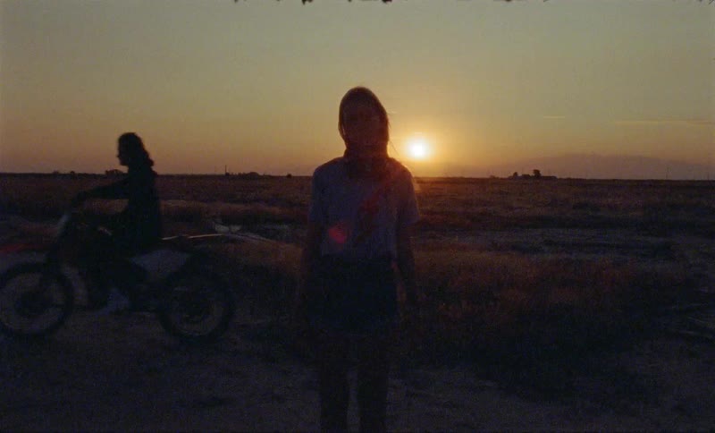 A young woman stands outdoors as the sun hangs very low in the sky and a man on a dirt back rides by behind her