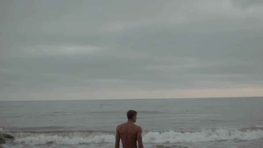 Shirtless muscular man looks at the sea on the sand beach on a cloudy day