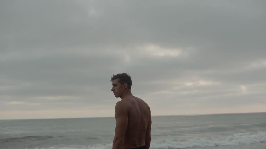 A muscular young man stands on a beach facing the ocean and waves but looking intensely over his shoulder