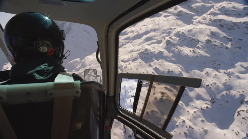 A pilot flying a helicopter over snow covered slopes on a sunny day