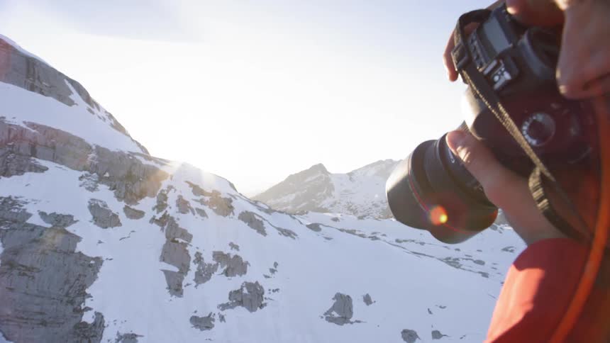 A photographer sits on the edge of a helicopter that is flying over a mountain range