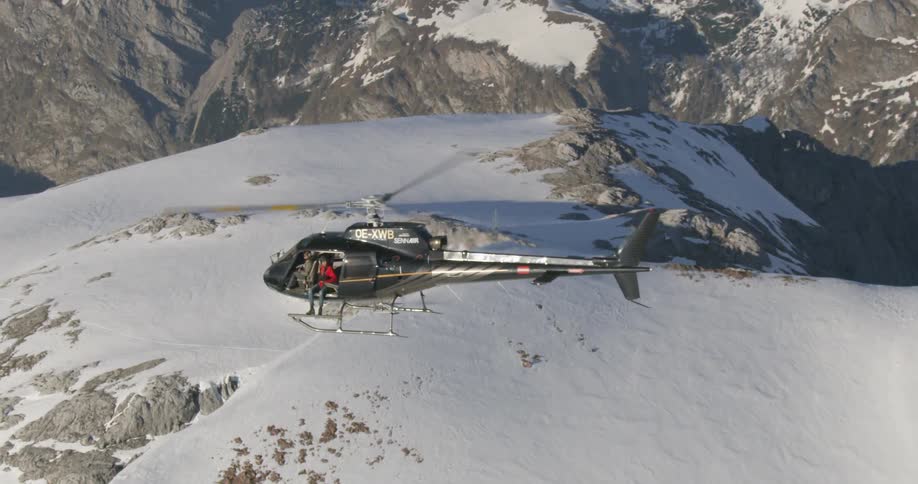 A man sits on the edge of a helicopter as if flies over the snowy mountains