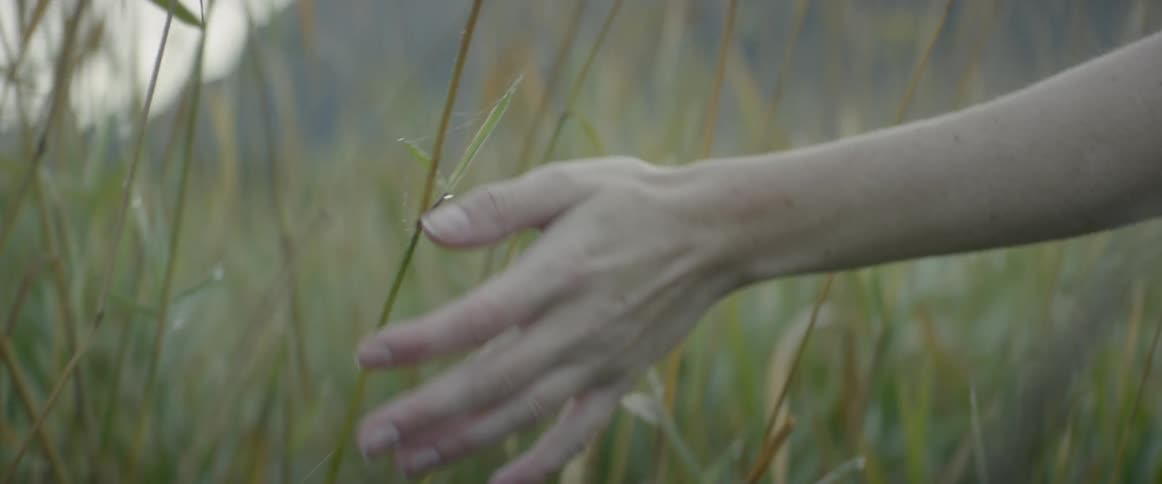 A white woman walks down the side of a road and brushes weeds with her hand
