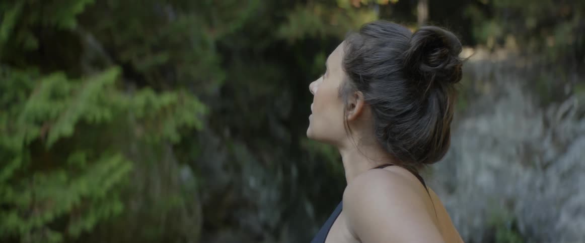 A woman is outdoors and surrounded by a thick grove of trees as she looks upwards at them