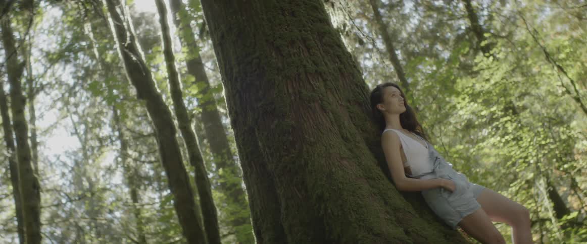 A woman leans on a tree in wooded area