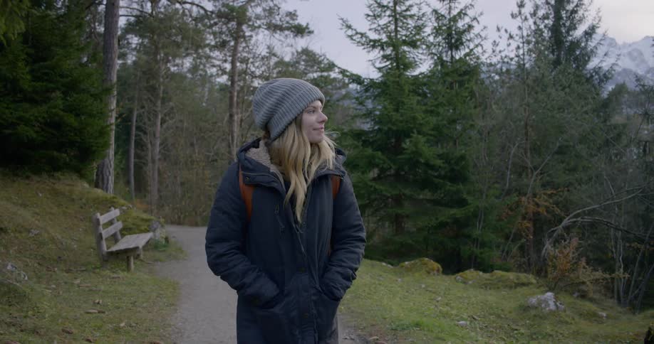 A white female dressed for cold weather and carrying a backpack is walking slowly on a paved path in a park with evergreens around her and mountains in the distance