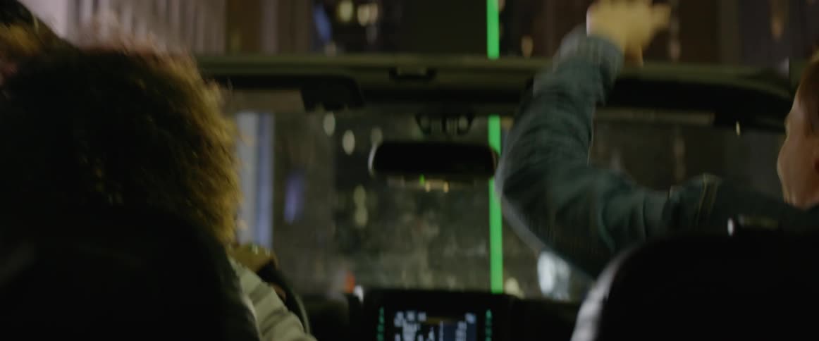A woman driving a car through a city while a man sits in the passenger seat pointing at things outside the car