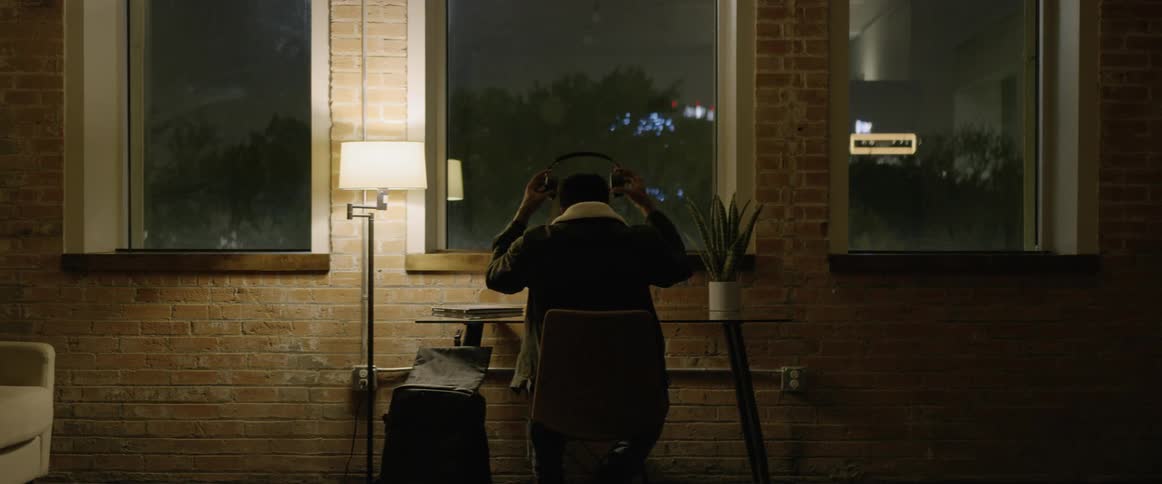 A black male is sitting in a chair at a desk in front of a wall with three large windows and puts on a pair of overtheear headphones