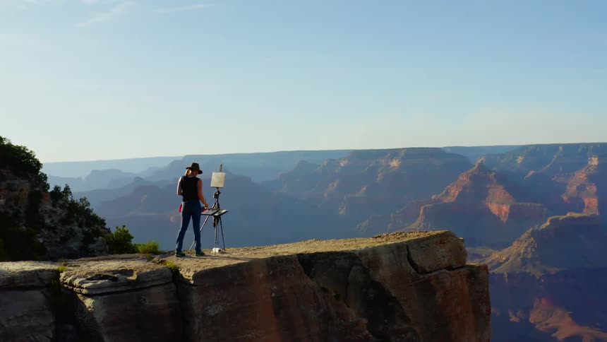A woman wearing a hat is painting onto a canvas held up by an easel atop a mountain