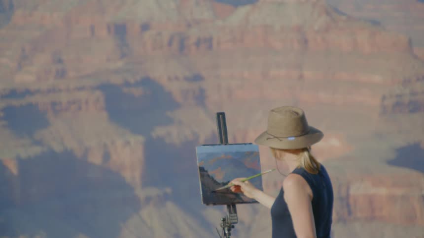 A woman wearing a hat is painting onto a canvas held up by an easel atop a mountain