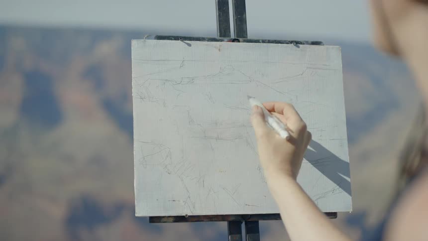 A woman wearing a hat is painting onto a canvas held up by an easel atop a mountain