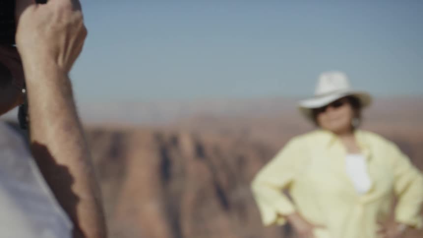 An elderly man takes a photo of a mature woman posing in front of a canyon