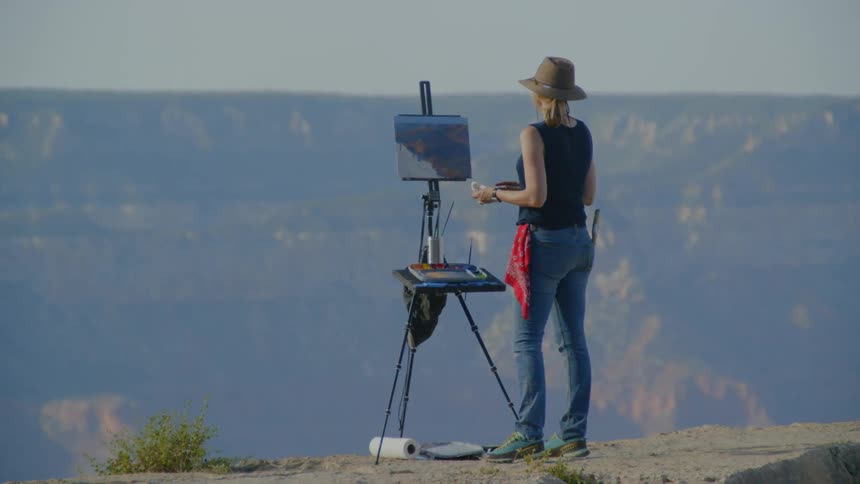 A woman wearing a hat and sunglasses painting onto a canvas held up by an easel atop a mountain