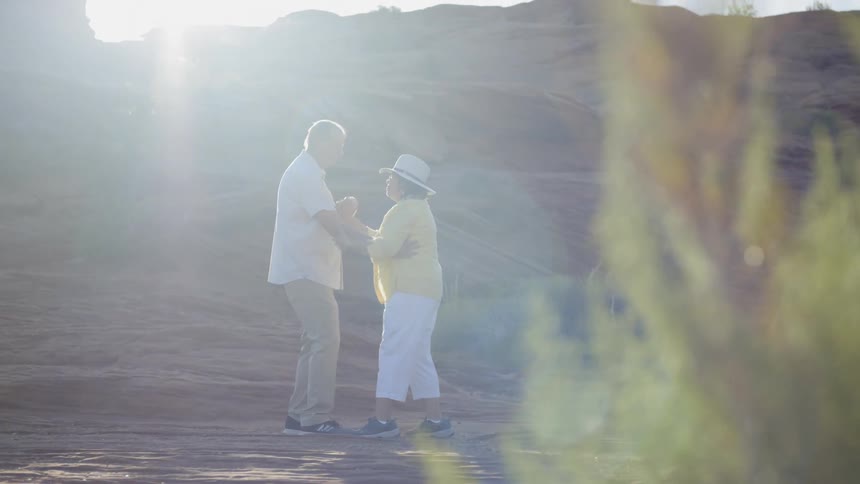 An older man and woman stand outside and dance together underneath the bright sunlight