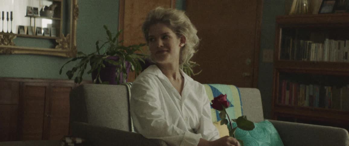 Blonde woman sitting on the couch holds a rose and smiles indoors during daylight