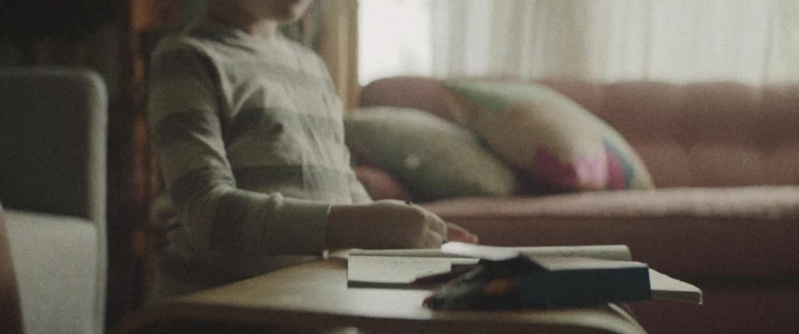 A young male child colors on a coloring book with a crayon in the living room while his mother reads