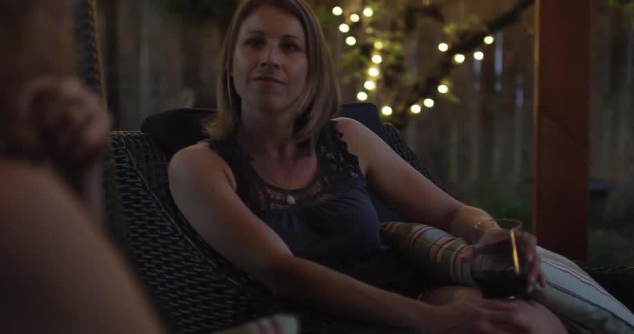 Blonde woman sitting in a wicker chair and holding a glass of wine as another blonde woman speaks