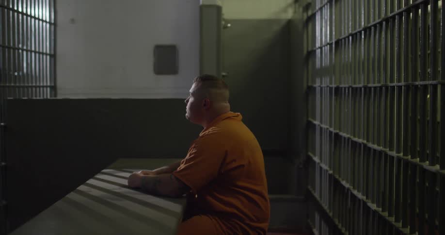 Male prisoner sits behind bars at a table, looking around himself