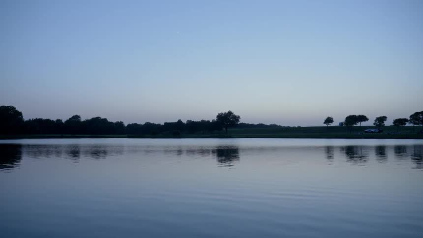 A tranquil lake has subtle ripples across its surface as a silhouette of the trees and bushes that line it can be seen