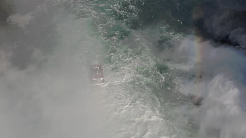 Water rushes down the side of a cliff into a river as mist rises around a boat