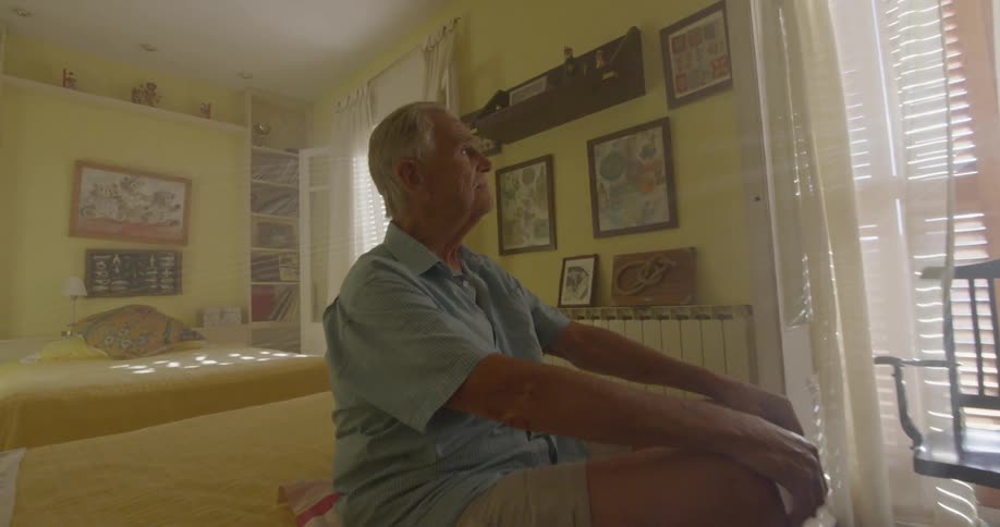 Man sits up straight on edge of bed looking toward window
