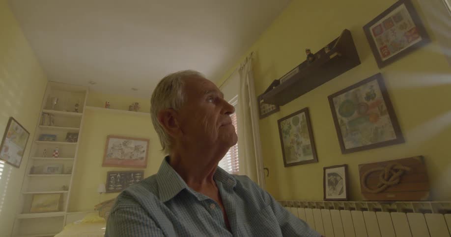 An elderly man appears to be sad while looking out a window in a room with walls painted yellow and covered in framed pieces of art