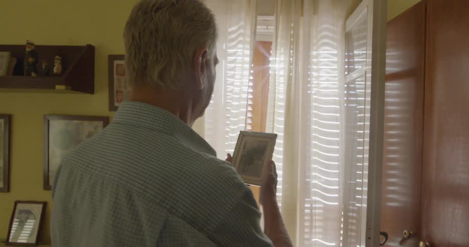 Elderly man walking over to window to look at framed picture he is holding
