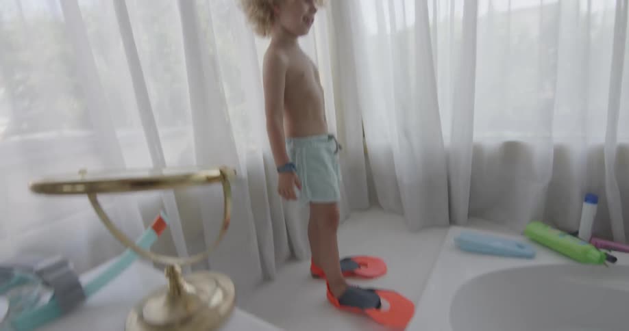 Little boy with blonde curly hair wearing flippers standing at edge of bathtub