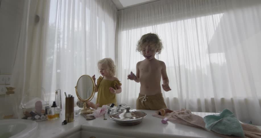 Two young children are in a bathroom and playing with makeup while looking into a mirror
