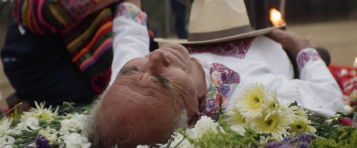 A elderly man with his eyes closed is on his back atop a pile of flowers as a woman strokes his arm