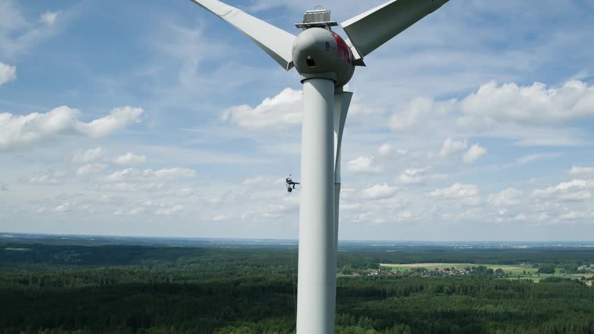 A person using a rope to descend a tall wind turbine that looms over a forest