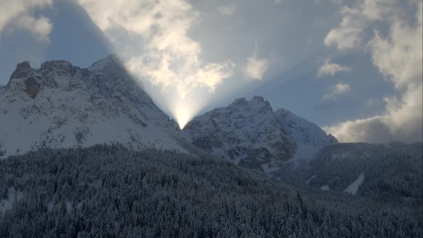 Sunlight shining between two mountain peaks above a pine forest
