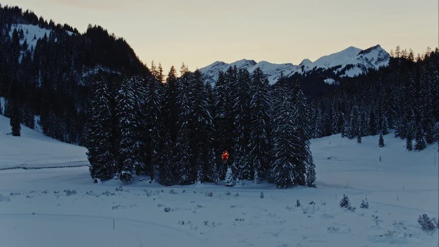 A small drone copter flies towards a dense forest in front of a mountain located in a snow covered landscape