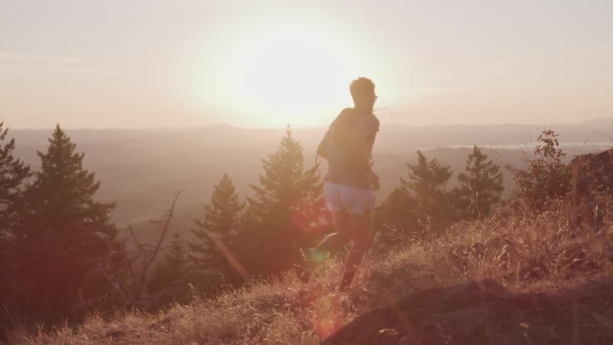 A man with a hydration backpack on is running up a hill with evergreens to his left and the sun on the horizon in the distance