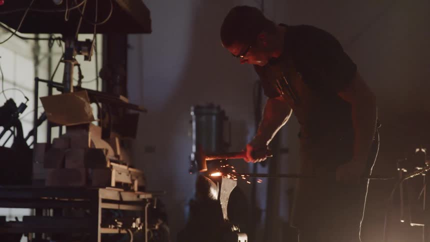 Blacksmith hammers on bar laying on anvil in workshop