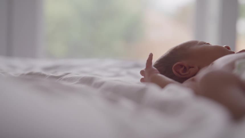 A small infant rests on its back atop a soft white blanket in front of an open window
