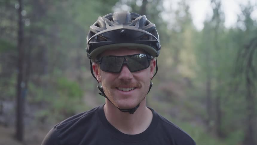 A man in a cycling helmet and dark sunglasses is smiling with tall trees in the background