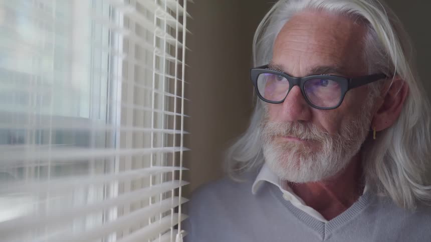 An elderly man with long white hair and glasses looks through the blinds over a window