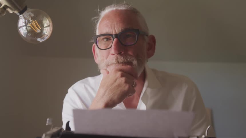 Senior bearded man sitting at the table turns to the camera in a room