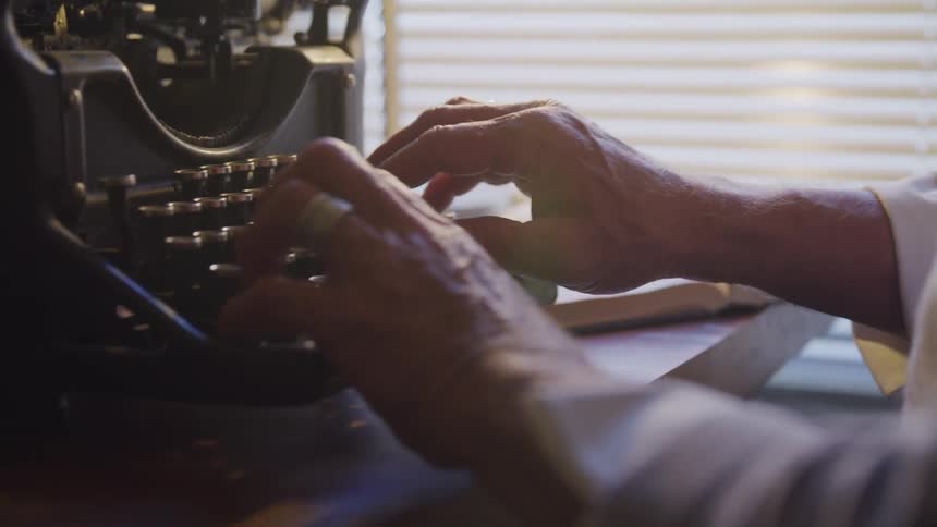 A man sitting at a desk by a window begins to type on an old fashioned type writer